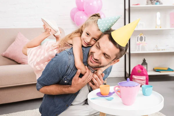 Père piggybackking fille en chapeau de cône tout en jouant thé partie à la maison — Photo de stock