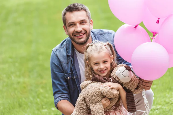 Батько і дочка з плюшевим ведмедем і рожевими кульками на газоні — стокове фото