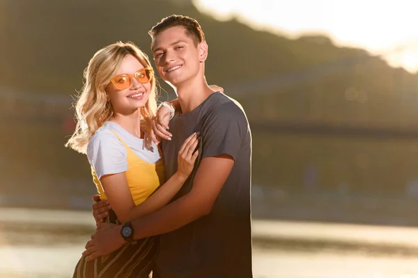 Smiling young couple hugging on river beach in evening and looking at camera — Stock Photo