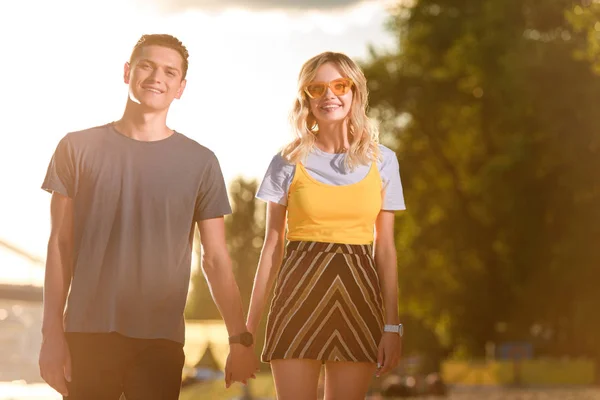 Smiling young couple holding hands on river beach in evening and looking at camera — Stock Photo