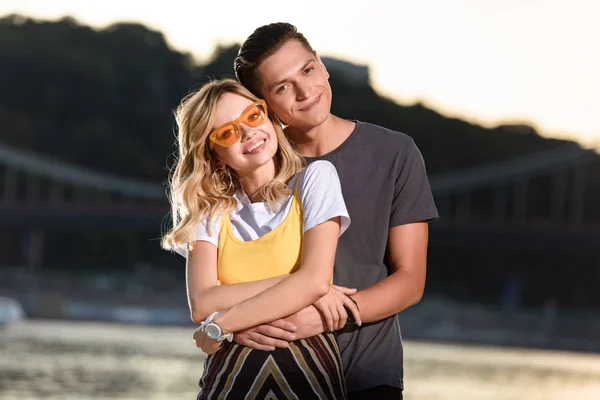 Boyfriend hugging girlfriend on river beach in evening and they looking at camera — Stock Photo