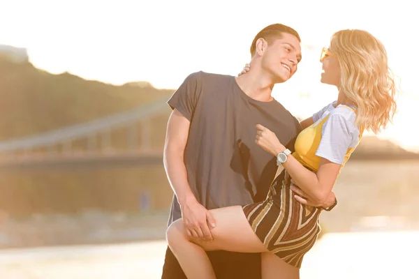 Jovem casal sorridente abraçando na praia do rio à noite e olhando um para o outro — Fotografia de Stock