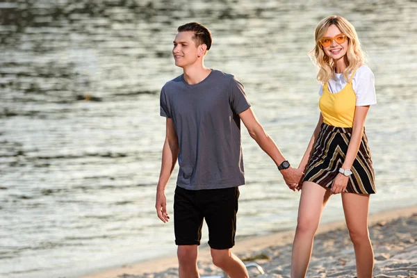 Pareja joven cogida de la mano y caminando por la playa del río por la noche - foto de stock