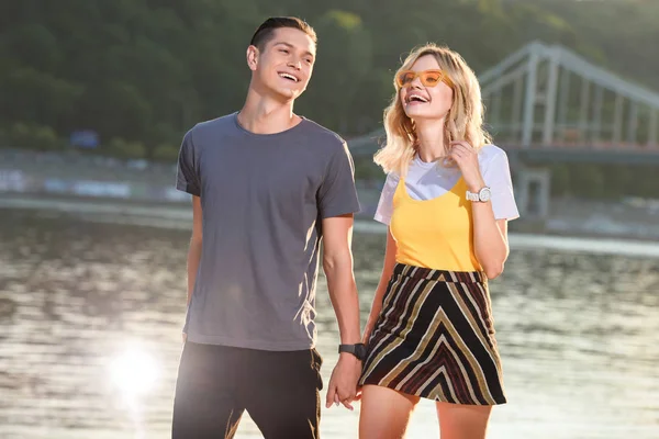 Laughing young couple holding hands and walking on river beach in evening — Stock Photo