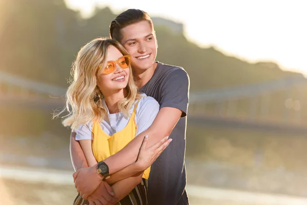 Smiling boyfriend hugging girlfriend on river beach in evening and they looking away — Stock Photo