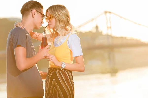 Side view of couple drinking beverage with two plastic straws from one bottle on river beach in evening — Stock Photo