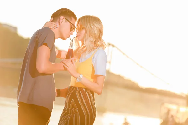 Couple drinking beverage with two plastic straws from one bottle on river beach during sunset — Stock Photo