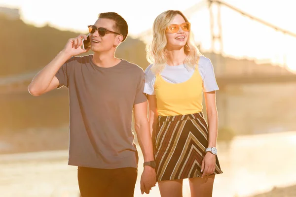 Giovane coppia che passeggia sulla spiaggia del fiume in serata, fidanzato che parla con lo smartphone — Foto stock
