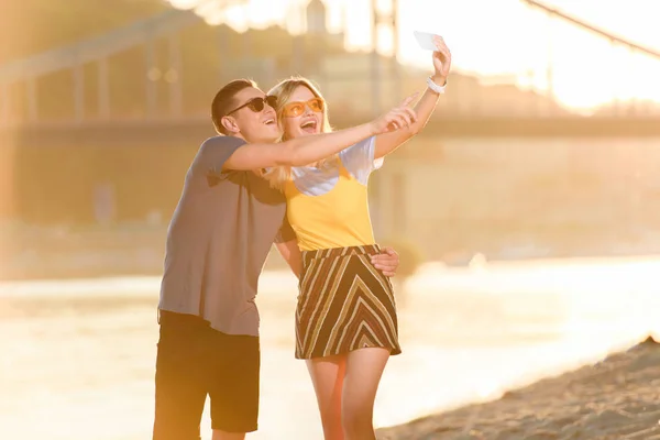 Jeune couple prenant selfie avec smartphone sur la plage de la rivière pendant le coucher du soleil — Photo de stock