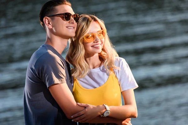 Молодая пара в солнечных очках обнимается на берегу реки вечером и смотрит в сторону — стоковое фото