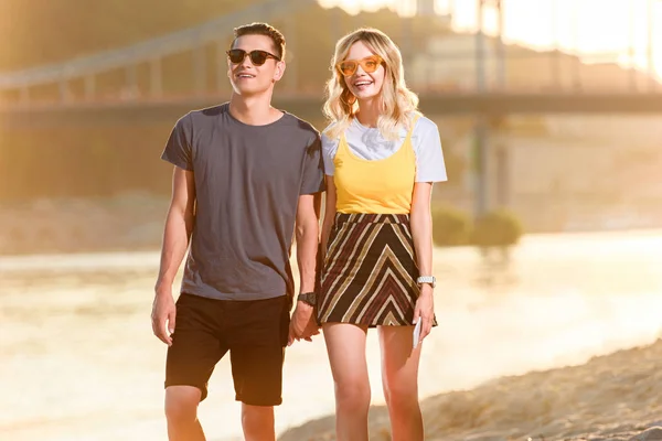 Junges Paar mit Sonnenbrille hält sich abends am Flussstrand an den Händen — Stockfoto