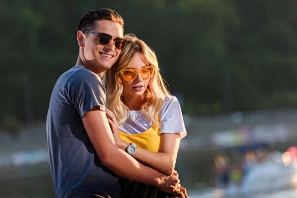 Boyfriend in sunglasses cuddling girlfriend on river beach in evening — Stock Photo