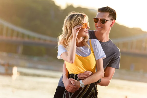 Fidanzato abbracciare fidanzata sulla spiaggia del fiume durante il tramonto, lei toccare occhiali da sole — Foto stock