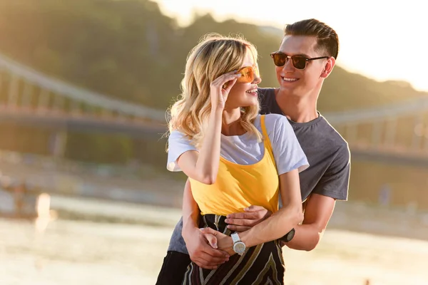 Copain étreignant petite amie sur la plage de la rivière pendant le coucher du soleil, elle touchant lunettes de soleil — Photo de stock