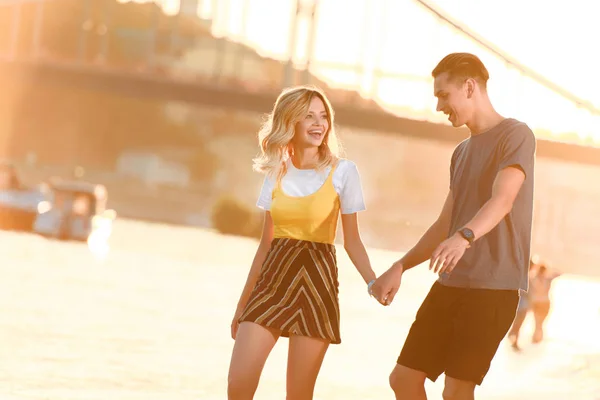 Усміхнена молода пара тримає руки і розважається на березі річки під час красивого заходу сонця — стокове фото