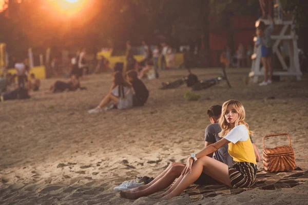 Jeune couple assis sur une couverture avec panier de paille sur la plage de la rivière le soir — Photo de stock