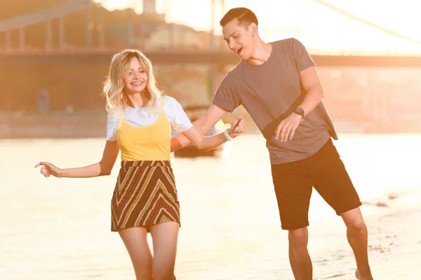 Riendo pareja joven divirtiéndose en la playa del río durante el atardecer - foto de stock