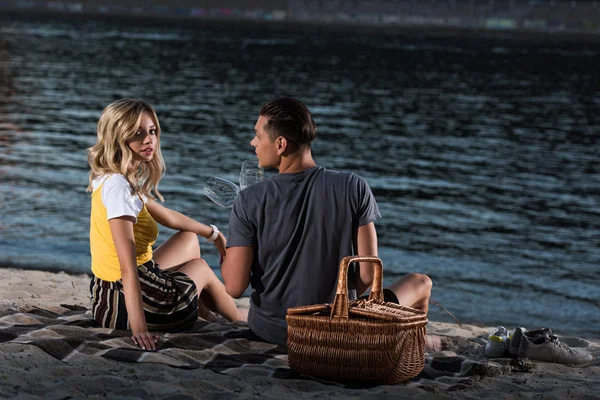 Jeune couple assis sur une couverture avec des verres à vin sur la plage de la rivière le soir — Photo de stock