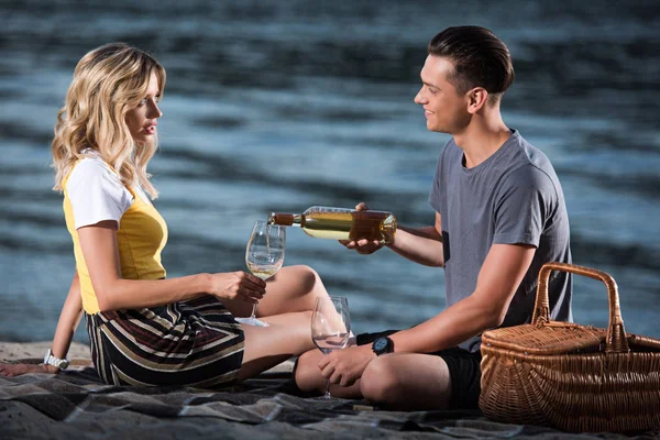 Side view of boyfriend pouring wine in glasses at picnic on river beach in evening — Stock Photo