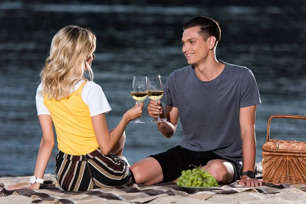 Seitenansicht eines jungen Paares, das bei einem Picknick am Flussstrand am Abend mit Weingläsern klingelt — Stockfoto