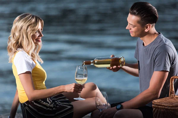 Copain joyeux verser du vin dans des verres au pique-nique sur la plage de la rivière en soirée — Photo de stock