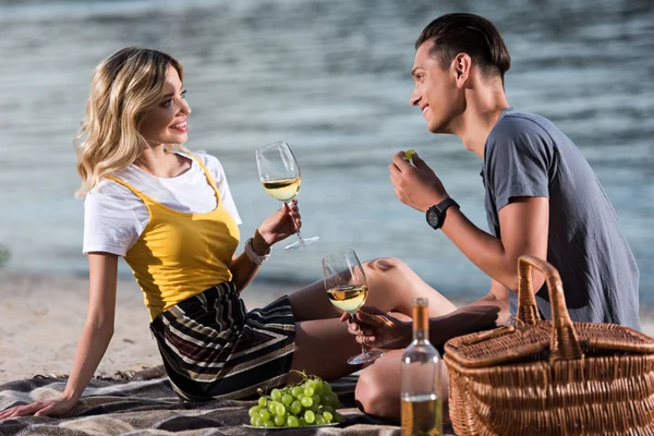 Smiling young couple drinking wine and eating grapes at picnic on river beach in evening — Stock Photo