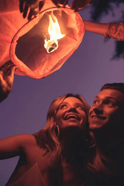 Vista de ángulo bajo de feliz pareja joven lanzando linterna del cielo en la playa del río por la noche - foto de stock