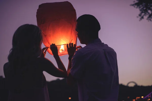 Vista trasera de la joven pareja lanzando linterna del cielo en la playa del río por la noche - foto de stock