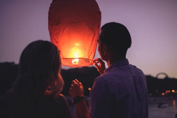Visão traseira do jovem casal lançando lanterna céu na praia do rio à noite — Fotografia de Stock