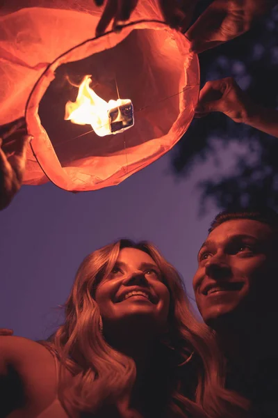 Baixo ângulo de visão de casal jovem sorrindo lançando lanterna céu na praia do rio à noite — Fotografia de Stock