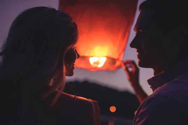 Портрет пары, запускающей небесный фонарь на берегу реки вечером и смотрящей друг на друга — стоковое фото