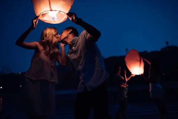 Jeune couple baisers et tenant ciel lanterne sur la plage de la rivière dans la soirée — Photo de stock