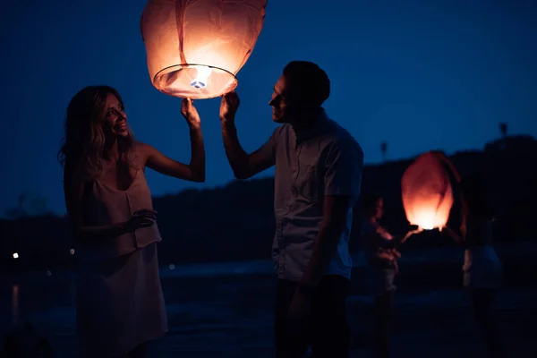 Feliz joven pareja lanzando linterna de cielo en la playa del río por la noche - foto de stock