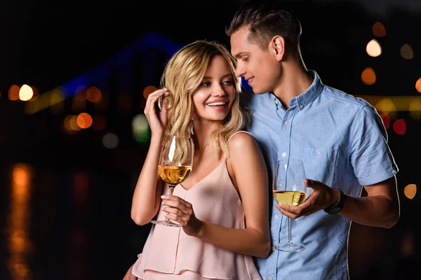 Heureux jeune couple debout avec des verres de vin blanc sur la plage de la rivière en soirée — Photo de stock