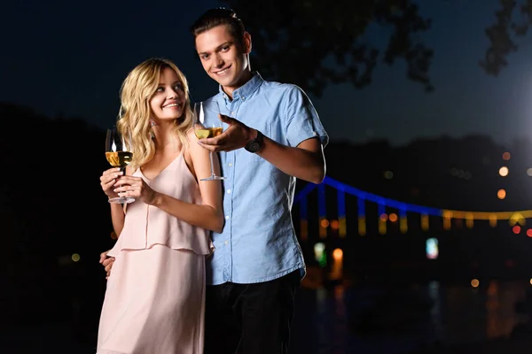 Sonriente joven pareja de pie con copas de vino en la playa del río por la noche - foto de stock