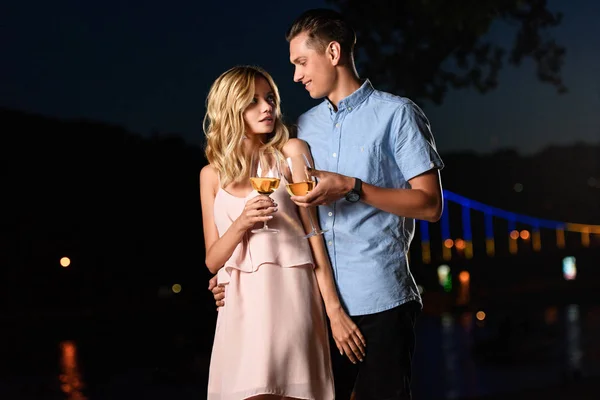 Young couple clinking with glasses of wine and looking at each other on river beach in evening — Stock Photo