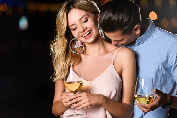 Boyfriend kissing smiling girlfriends shoulder and they holding glasses of wine in evening — Stock Photo