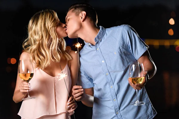 Casal beijando e segurando copos de vinho e faíscas de festa à noite — Fotografia de Stock