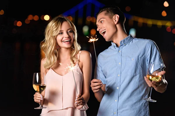 Rire jeune couple tenant des verres de vin et des mousseux de fête sur la plage de la rivière dans la soirée — Photo de stock