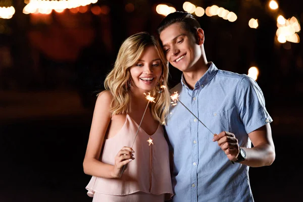 Happy young couple holding party sparkles on river beach in evening — Stock Photo