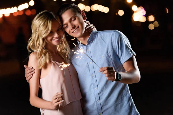 Sonriente joven pareja celebración de fiesta brilla en la playa del río por la noche - foto de stock