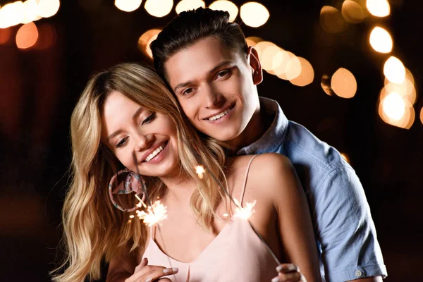 Happy young couple hugging and holding party sparkles on river beach in evening — Stock Photo