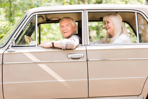 Vista lateral de la sonriente pareja de ancianos sentados en un coche vintage beige - foto de stock