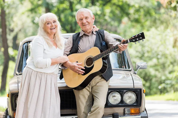 Seniorin umarmt Mann mit Gitarre gegen beiges Auto — Stockfoto