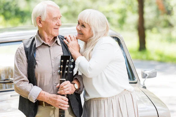 Marido y mujer mayores mirándose el uno al otro y el hombre sosteniendo la guitarra apoyada en el coche - foto de stock