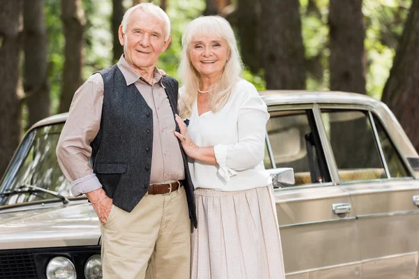 Pareja de ancianos de pie cerca de coche vintage y mirando a la cámara - foto de stock