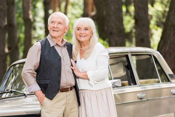 Pareja de ancianos de pie cerca de coche retro y mirando hacia otro lado - foto de stock