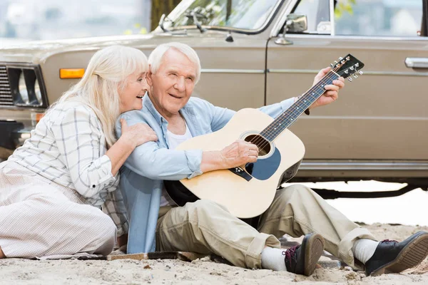 Seniorin sitzt mit Mann auf Sand und spielt Gitarre gegen Retro-Auto — Stockfoto