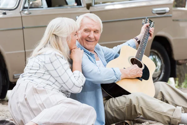 Senior wife sitting on sand with husband playing guitar against beige car — Stock Photo
