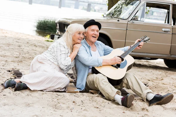 Femme âgée souriante assise sur le sable avec l'homme jouant de la guitare contre la voiture beige — Photo de stock